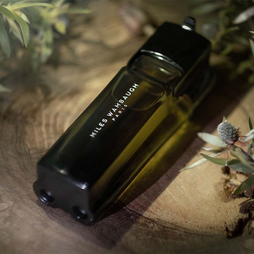 Elegant parfumflesje van Miles Wambaugh op een houten tafel, omringd door subtiele bloemelementen.