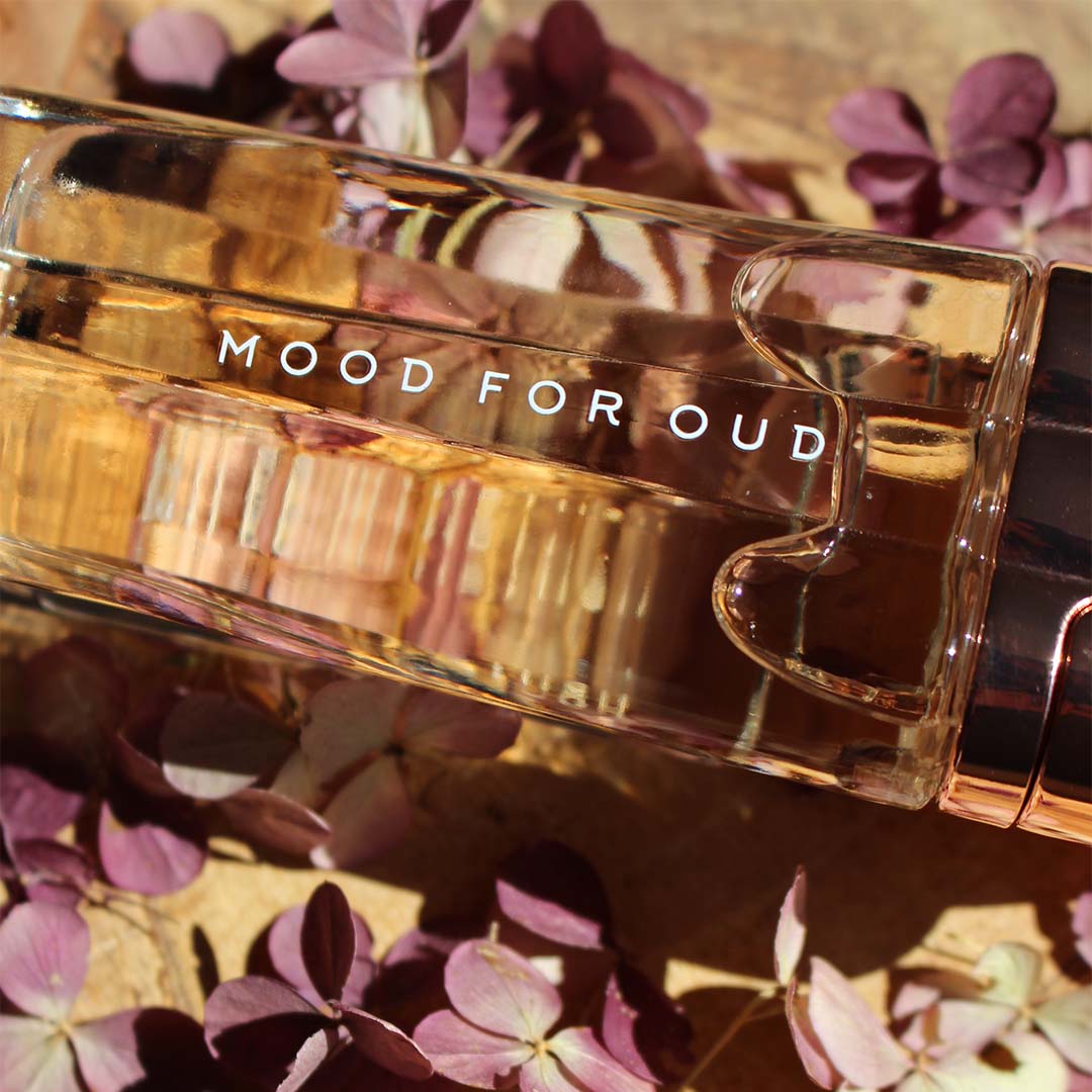 Elegant parfumflesje "Mood for Oud" van Miles Wambaugh liggend op een houten ondergrond, omringd door paarse bloemen.