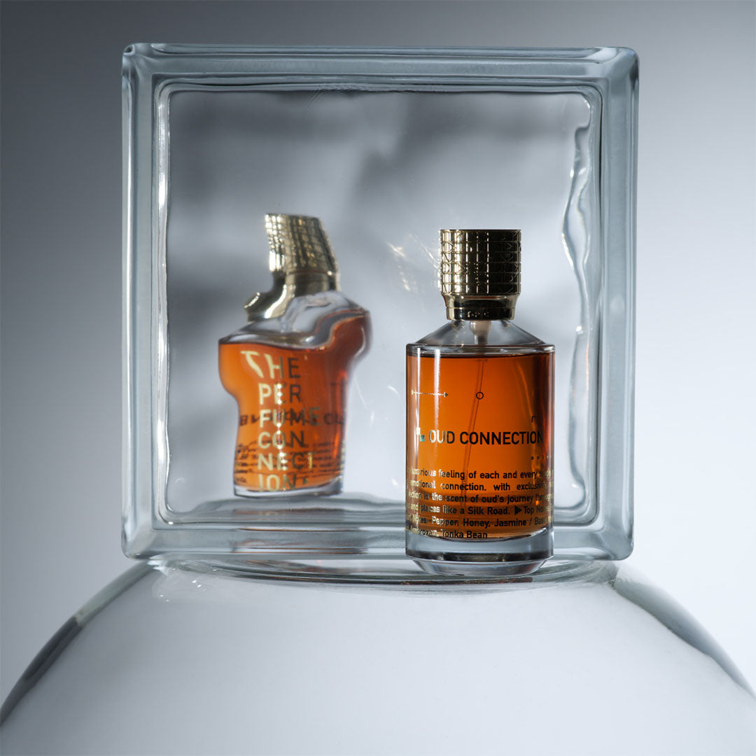 Parfumflesje "The Oud Connection" van The Perfume Connection met een reflectie in een glazen blok, tegen een grijze achtergrond.